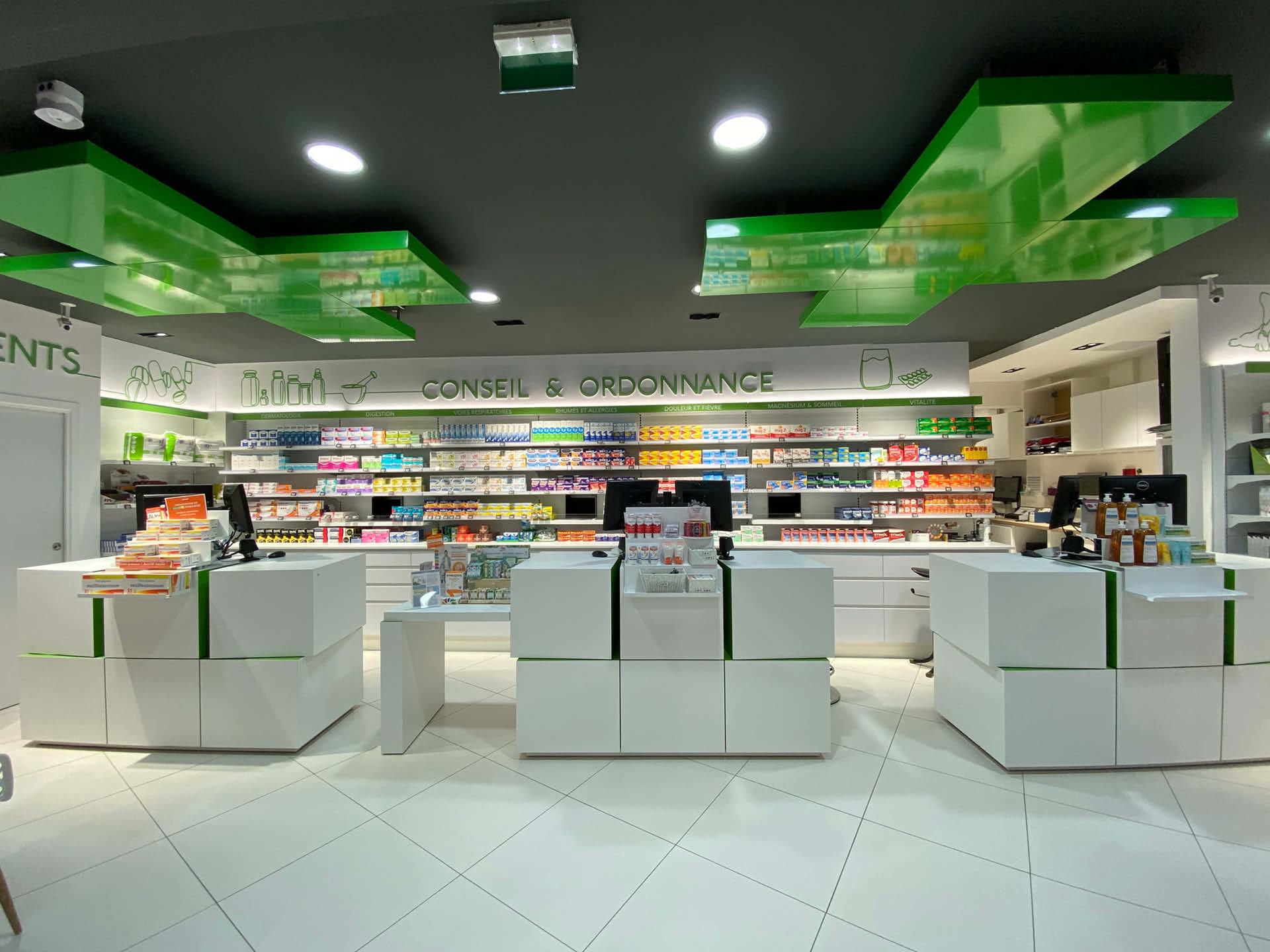 Comptoirs blancs lumineux sous un plafond gris marqué par la croix verte de la pharmacie, symbole du nouvel agencement moderne et accueillant de l’officine.