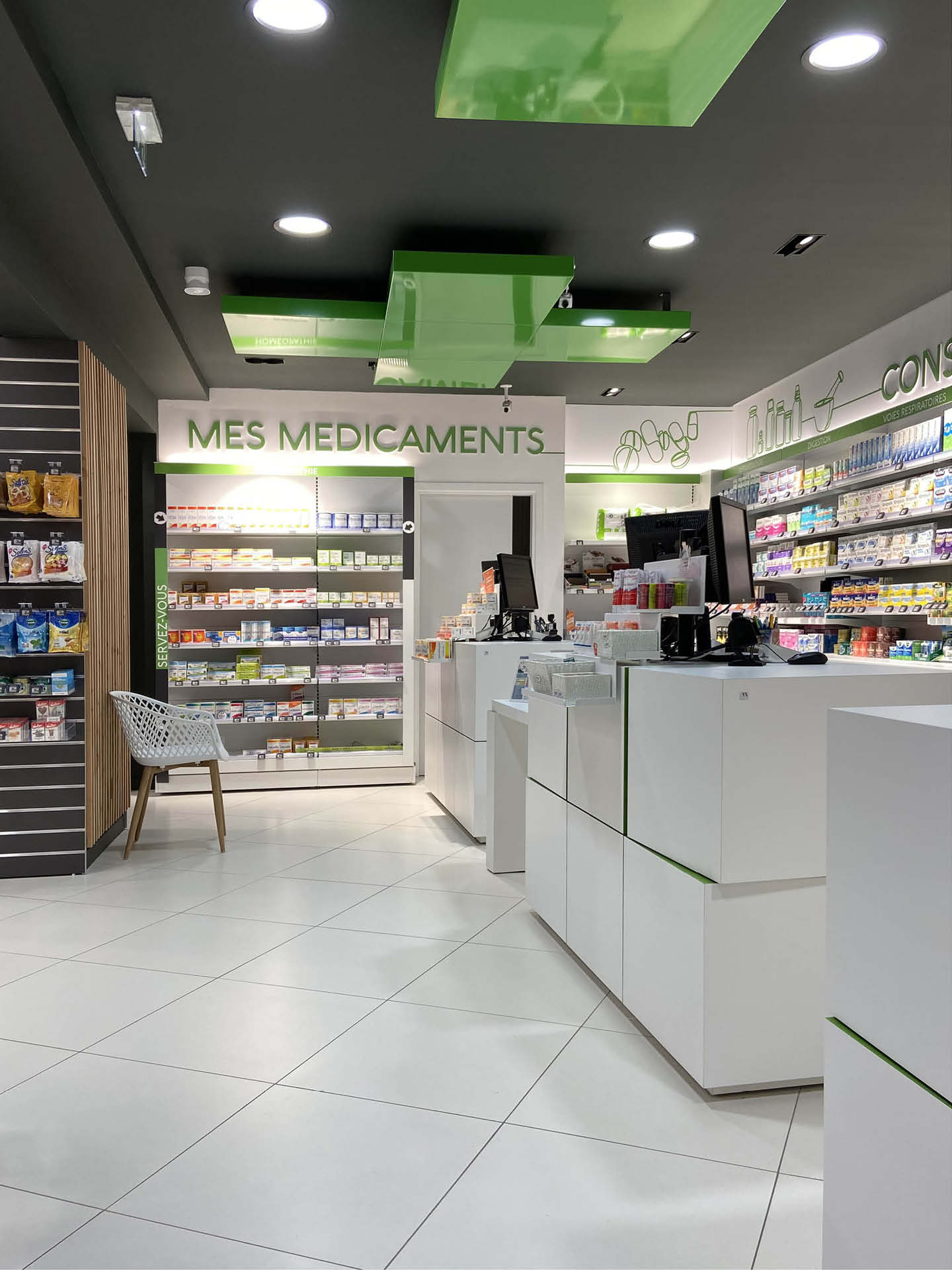 Comptoirs blancs modernes avec plafond gris et croix verte rétroéclairée, symbolisant l’identité de la Pharmacie Escarra et la modernité de son nouvel agencement.