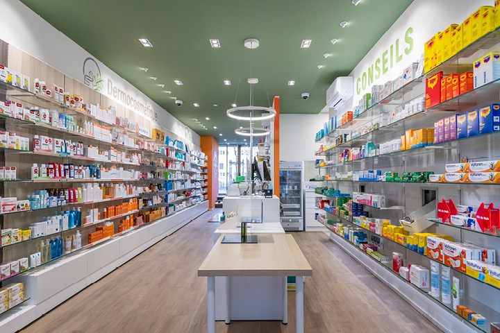 Vue d’ensemble de l’espace de vente de la Pharmacie Gare Chêne-Bourg, agencé en longueur avec des tons bois, vert et orange pour une ambiance chaleureuse.