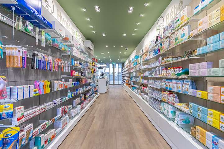 Vue d’ensemble de l’espace de vente de la Pharmacie Gare Chêne-Bourg, agencé en longueur avec des tons bois, vert et orange pour une ambiance chaleureuse.
