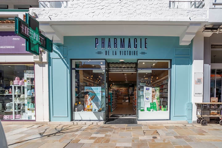 Façade rénovée de la Pharmacie de la Victoire avec couleurs bleu pastel, moulures en bois et grandes vitrines.