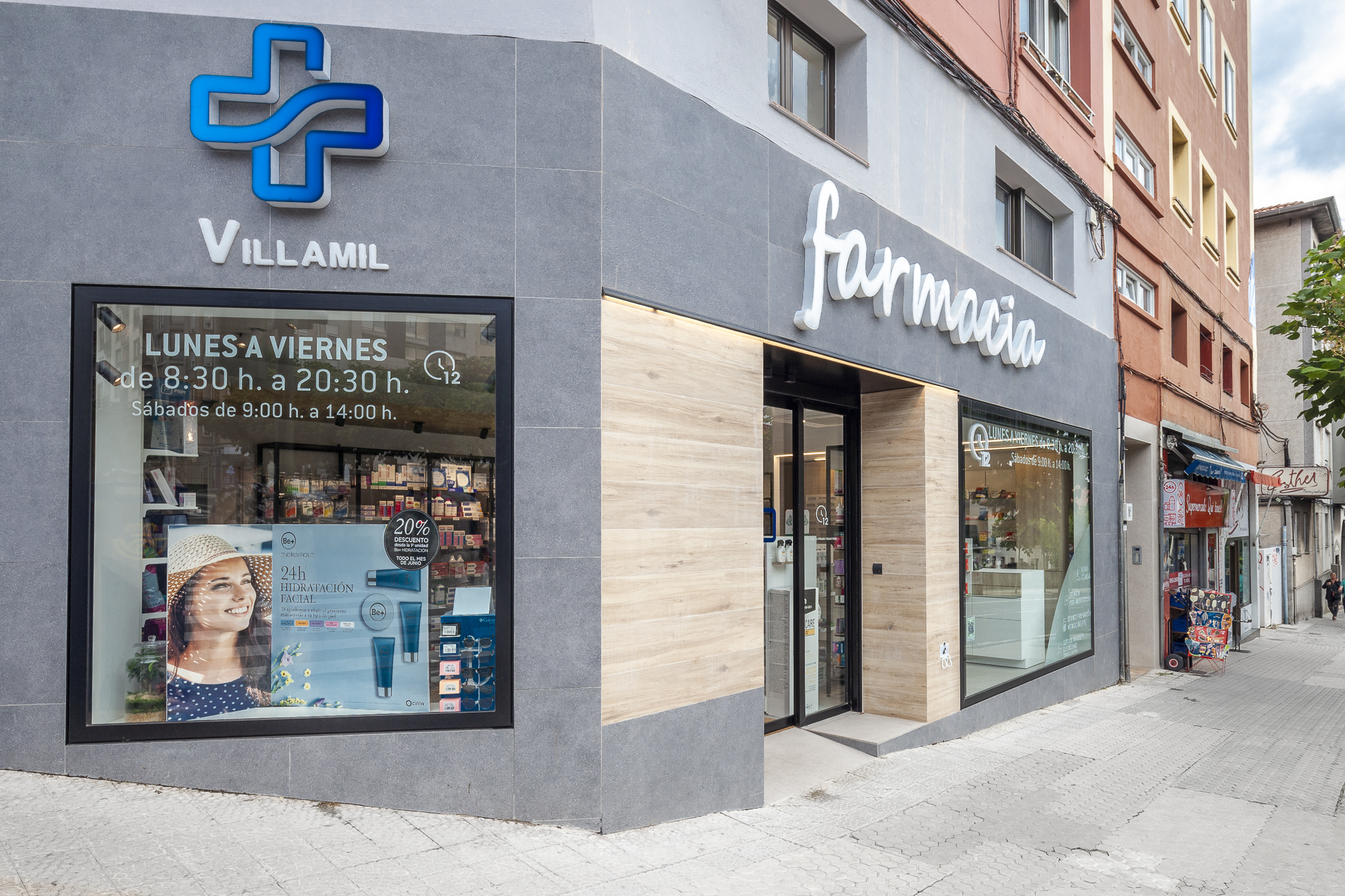 Fachada en esquina de la Farmacia Villamil en Santander, con revestimiento de madera, piedra gris y rótulo luminoso.