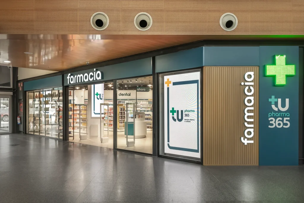Fachada de farmacia Segovia en centro comercial con revestimiento de listones de madera y rótulo de cruz verde.