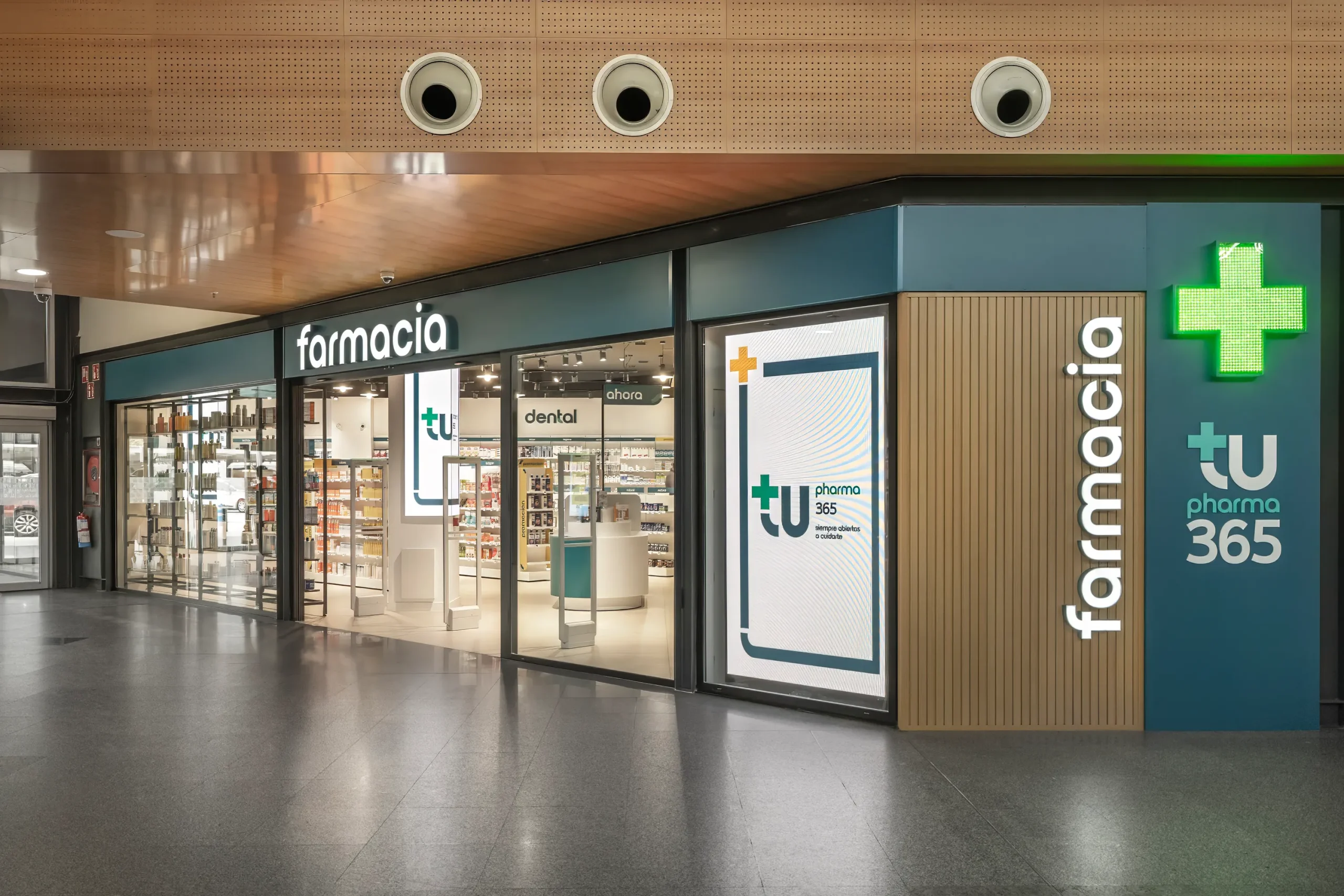 Fachada de farmacia Segovia en centro comercial con revestimiento de listones de madera y rótulo de cruz verde.