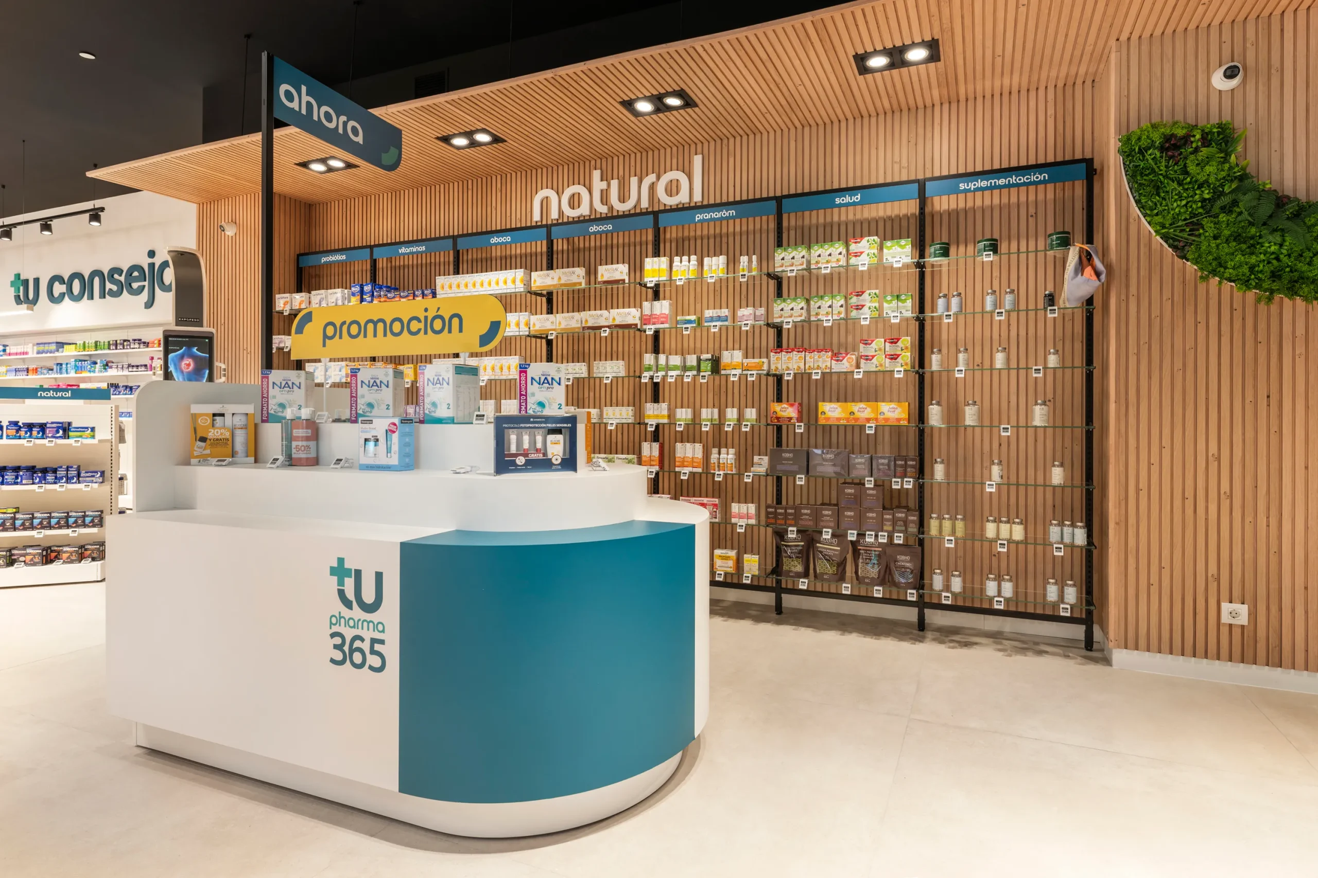 Mueble isla promocional turquesa en primer plano con área de parafarmacia natural al fondo.
