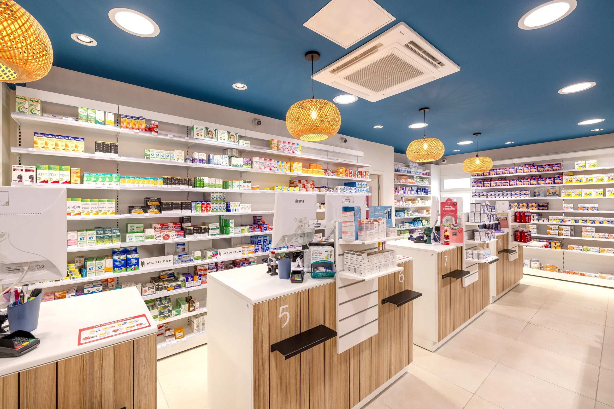 Comptoirs en bois et blanc de la Pharmacie du Golfe sous un plafond bleu et des suspensions en osier.