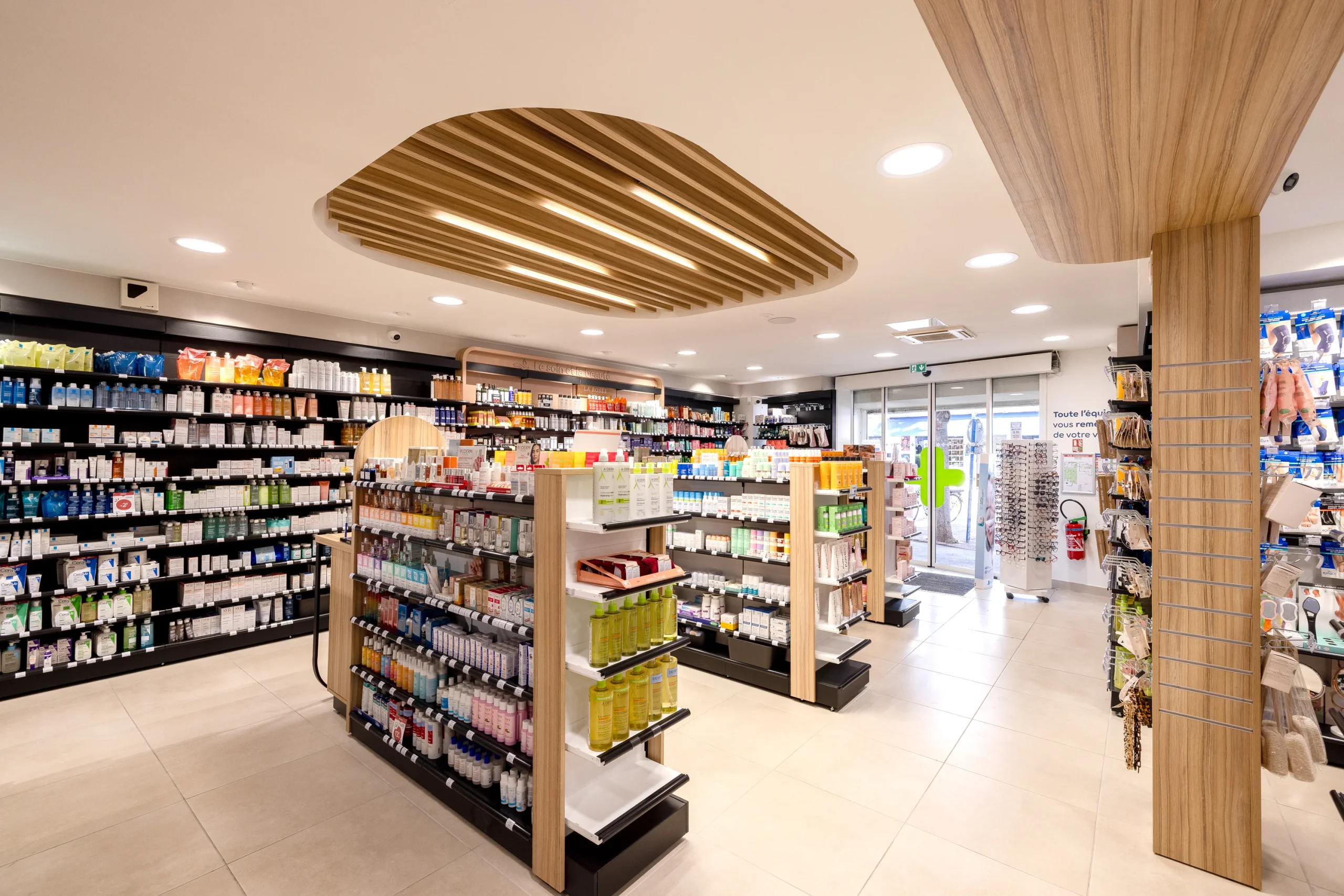 Gondoles sur-mesure de la Pharmacie du Golfe sous un plafond bois, avec vue sur l’entrée vitrée.
